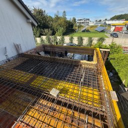 Zbrojenie stropu budynku w trakcie budowy, widok z góry. Żółte szalunki, stalowe pręty zbrojeniowe tworzą siatkę. W tle budynki, zieleń i błękitne niebo.