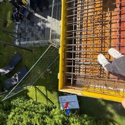 BUDOWLANIEC POMORZE SPÓŁKA Z OGRANICZONĄ ODPOWIEDZIALNOŚCIĄ - Z góry: szalunek zbrojony stalą na fundamencie, obok trawnik z krzewami, narzędzia i sprzęt budowlany. Widoczne stopy pracownika w białych butach.