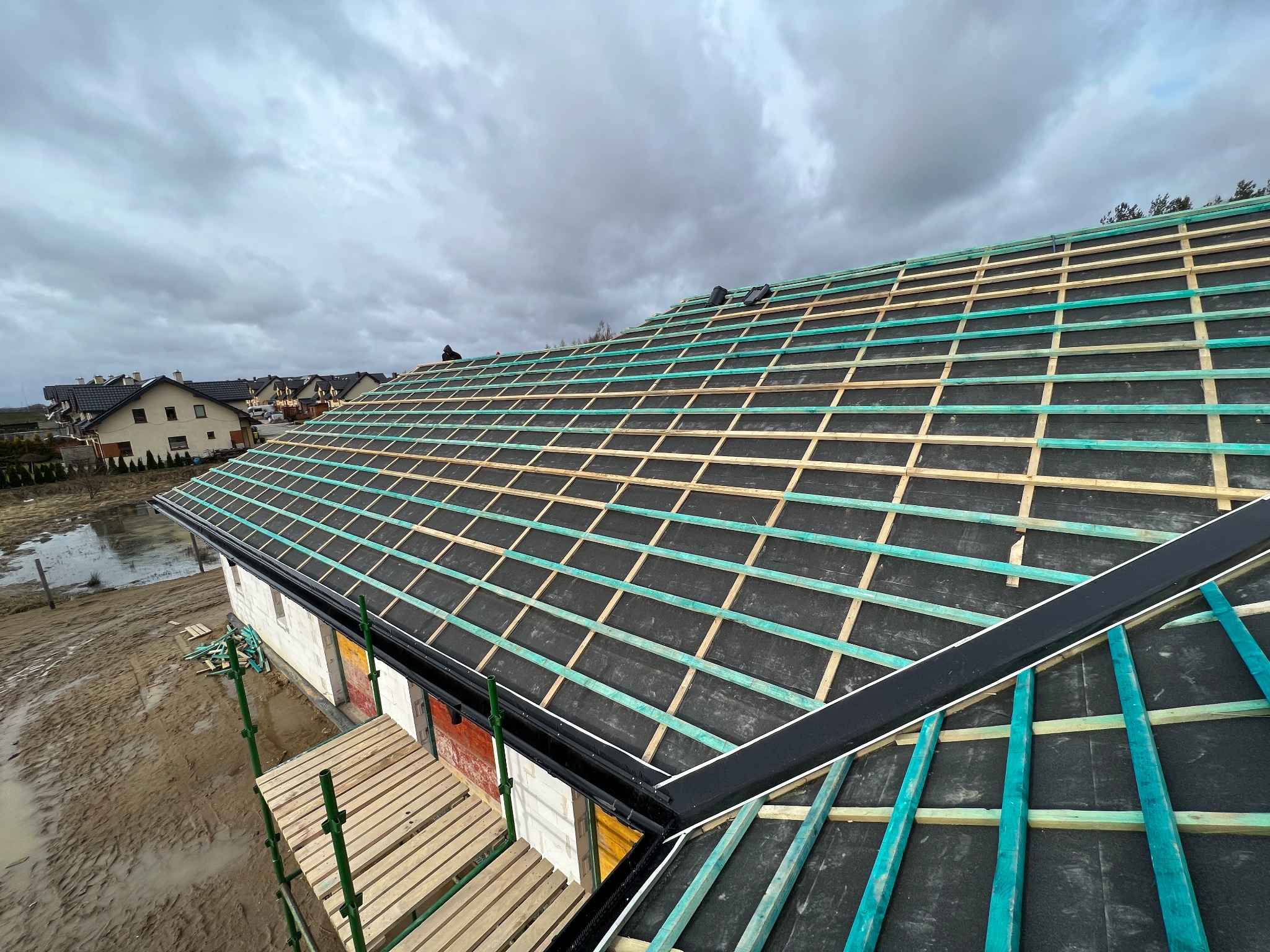 Konstrukcja dachu z łatami i kontrłatami na czarnej membranie, widok z góry na budynek w trakcie budowy, w tle zabudowa jednorodzinna i pochmurne niebo.