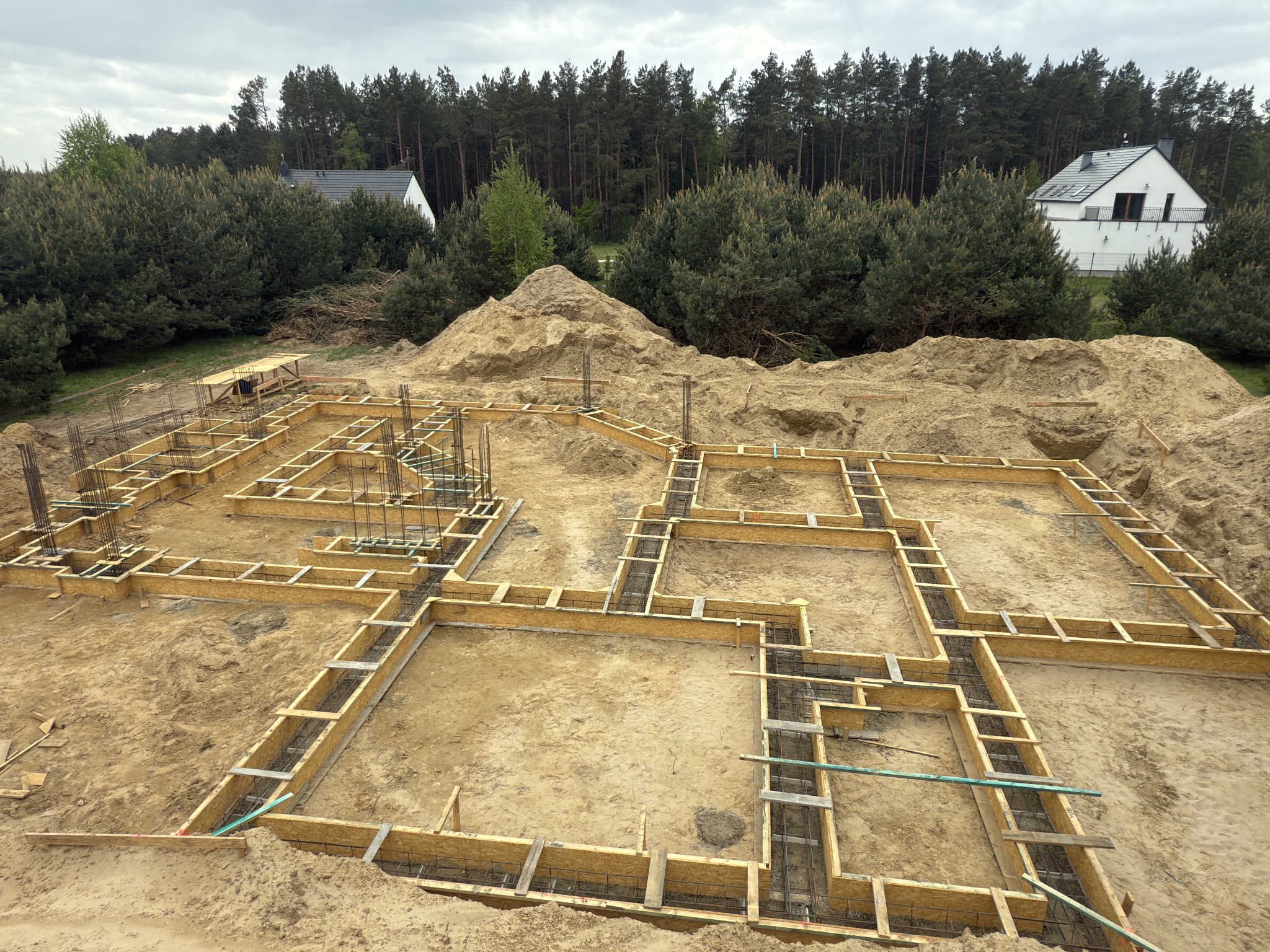 Szeroki kadr na szalunek fundamentów domu jednorodzinnego w piasku, z widocznym zbrojeniem i lasem w tle. Etap budowy z bliska, w dziennym świetle.