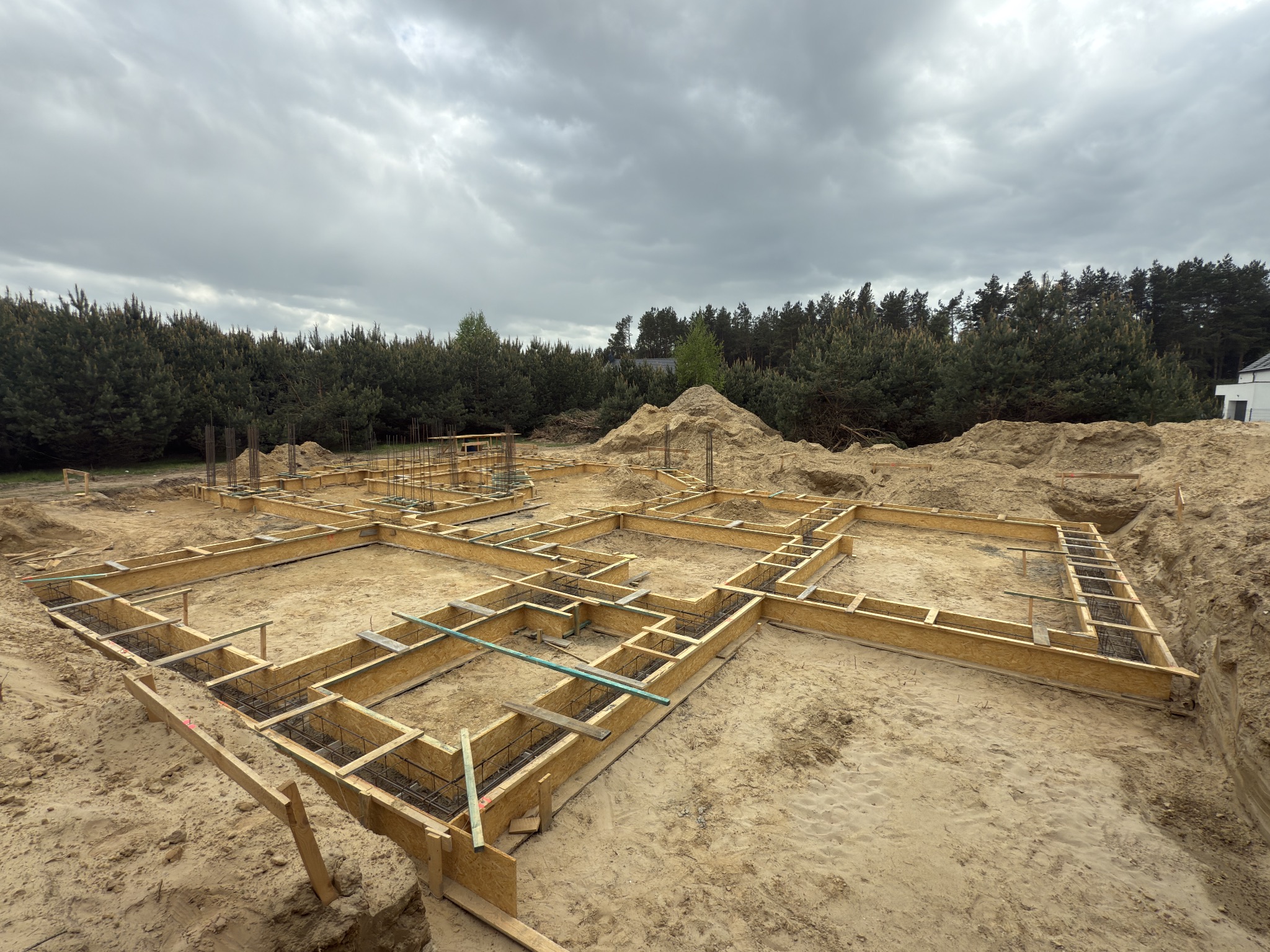 Szeroki kadr na szalunek fundamentów zbrojonych stalą, przygotowany do zalania betonem. W tle drzewa i pochmurne niebo. Widok z góry.