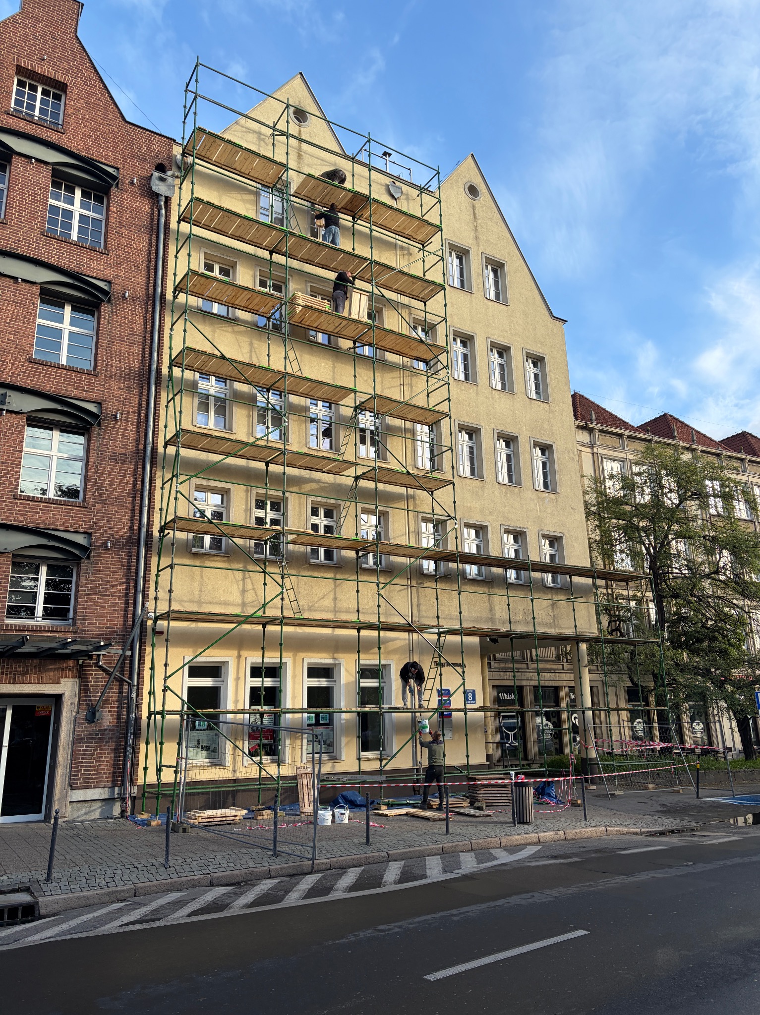 Żółta kamienica w trakcie renowacji, otoczona rusztowaniami. Pracownicy na różnych poziomach rusztowań. Widoczne detale architektoniczne i sąsiednie budynki.