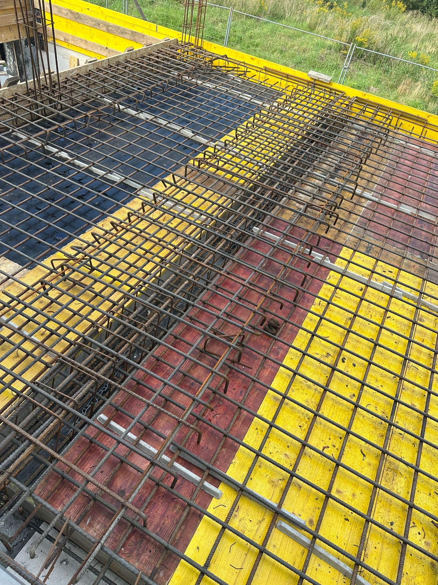 Zbrojenie stropu budynku w trakcie budowy, widok z góry. Żebrowana siatka stalowa na deskowaniu z żółtymi i czerwonymi elementami. Widoczne pręty pionowe.