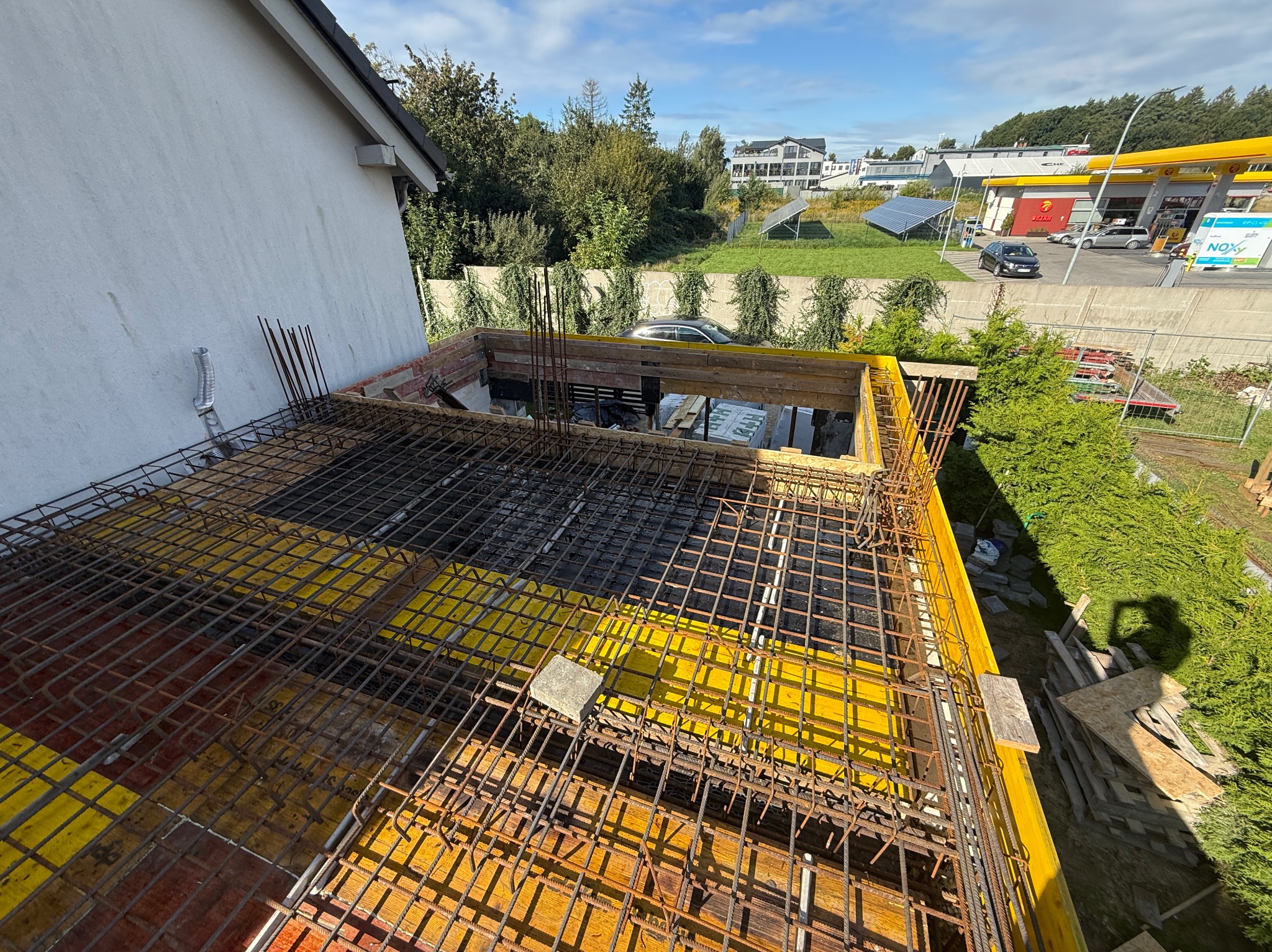 Zbrojenie stropu budynku w trakcie budowy, widok z góry. Żółte szalunki, stalowe pręty zbrojeniowe tworzą siatkę. W tle budynki, zieleń i błękitne niebo.