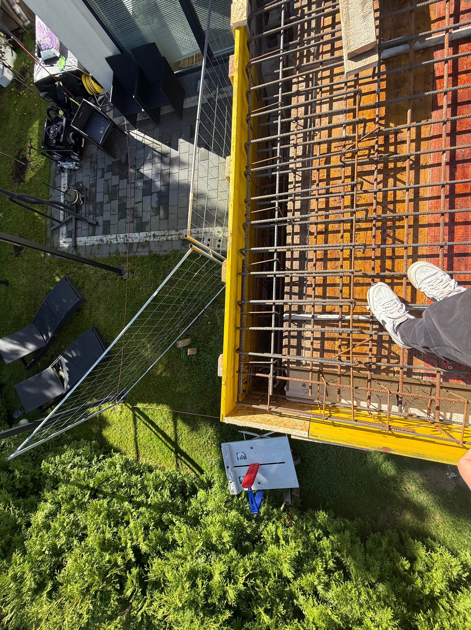 Z góry: szalunek zbrojony stalą na fundamencie, obok trawnik z krzewami, narzędzia i sprzęt budowlany. Widoczne stopy pracownika w białych butach.