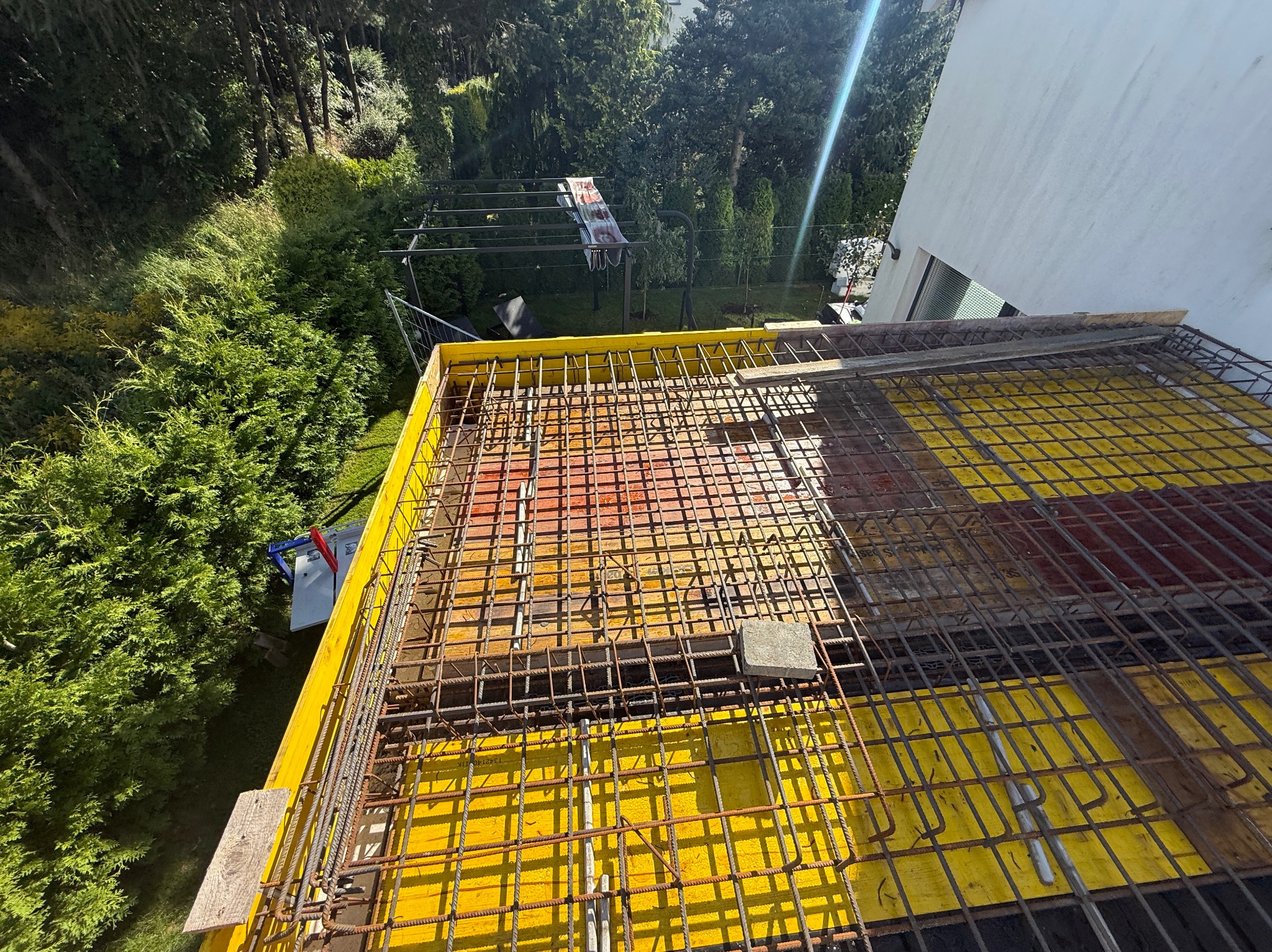 Żółte szalunki i zbrojenie na budowie domu, widok z góry. W tle zieleń drzew i krzewów, fragment białej ściany budynku. Konstrukcja przygotowana do zalania betonem.