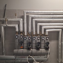 FUB Pulsar - Rozdzielacz instalacji grzewczej z pompami obiegowymi, rury zaizolowane folią aluminiową, na tle szarej ściany z płytek. Widoczne manometry i zawory.
