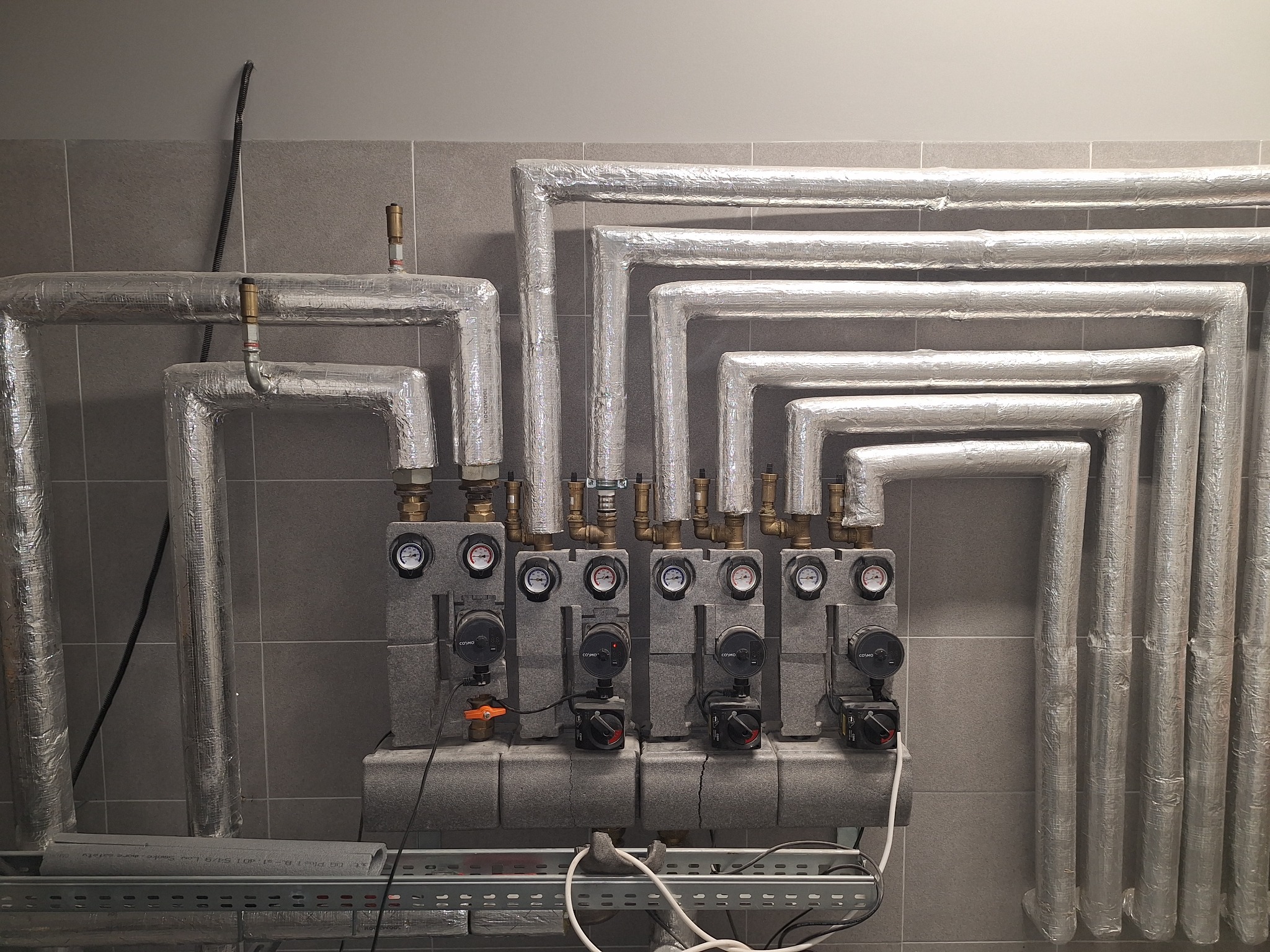Rozdzielacz instalacji grzewczej z pompami obiegowymi, rury zaizolowane folią aluminiową, na tle szarej ściany z płytek. Widoczne manometry i zawory.