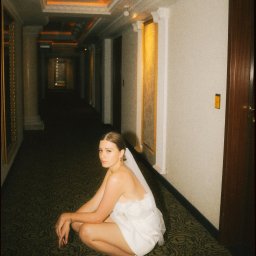 shutts.studio - Panna młoda w krótkiej sukience i welonie kuca w hotelowym korytarzu z wzorzystą wykładziną, spoglądając w obiektyw. Ciepłe oświetlenie i kolumny w tle.