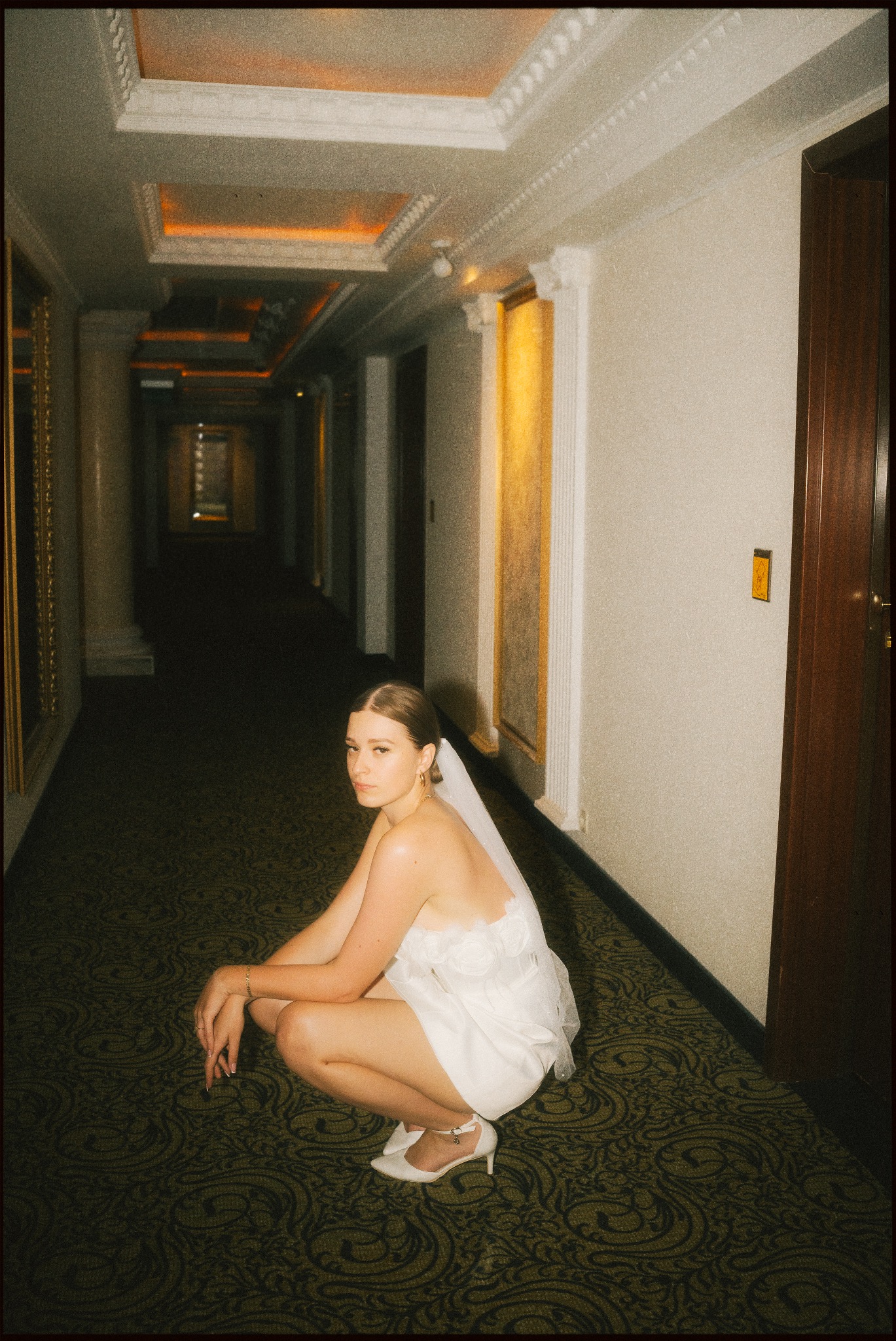 Panna młoda w krótkiej sukience i welonie kuca w hotelowym korytarzu z wzorzystą wykładziną, spoglądając w obiektyw. Ciepłe oświetlenie i kolumny w tle.