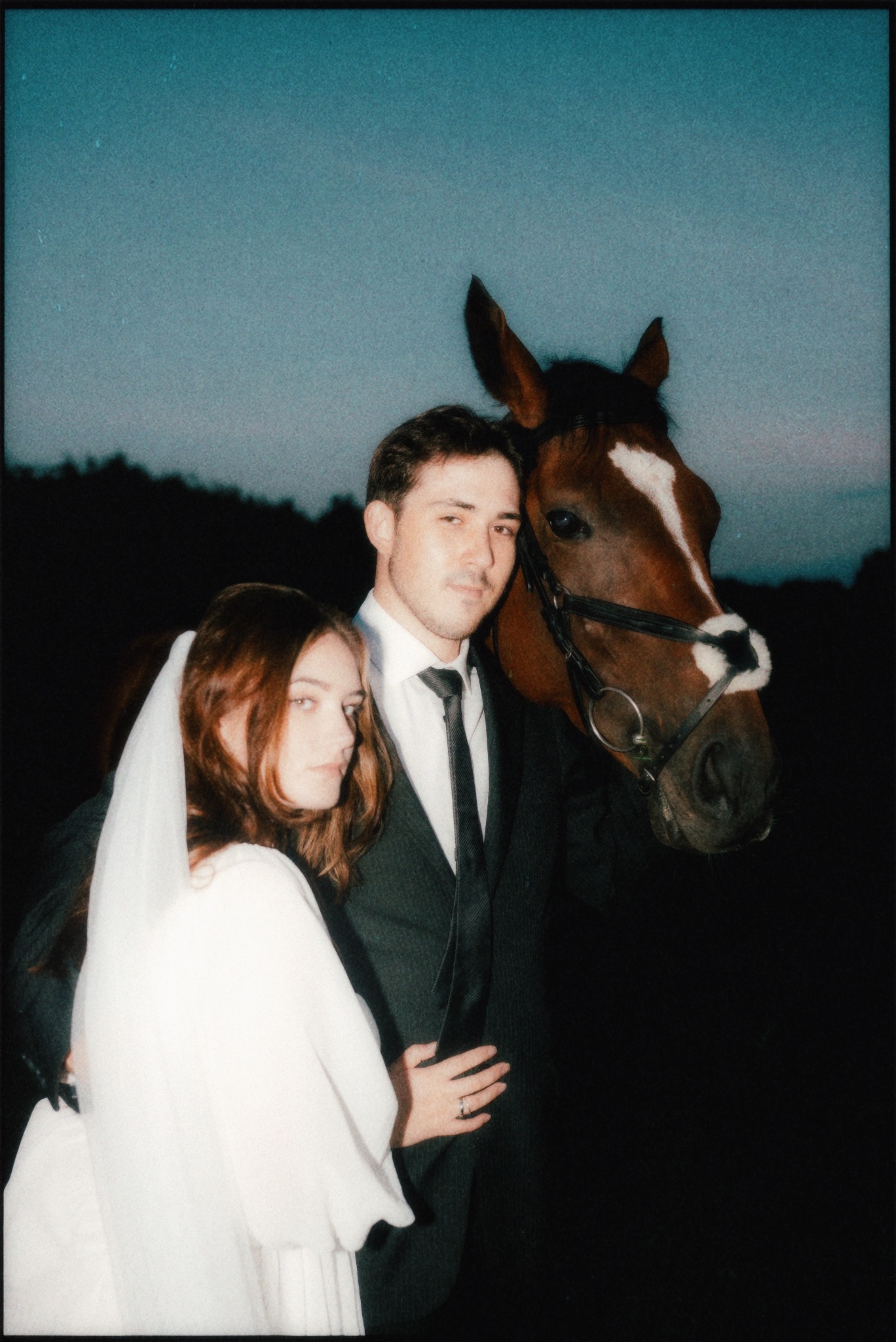 Para młoda w objęciach z koniem na tle wieczornego nieba. Ona w białej sukni i welonie, on w garniturze. Nietypowe ujęcie z sesji ślubnej, oryginalna pamiątka.
