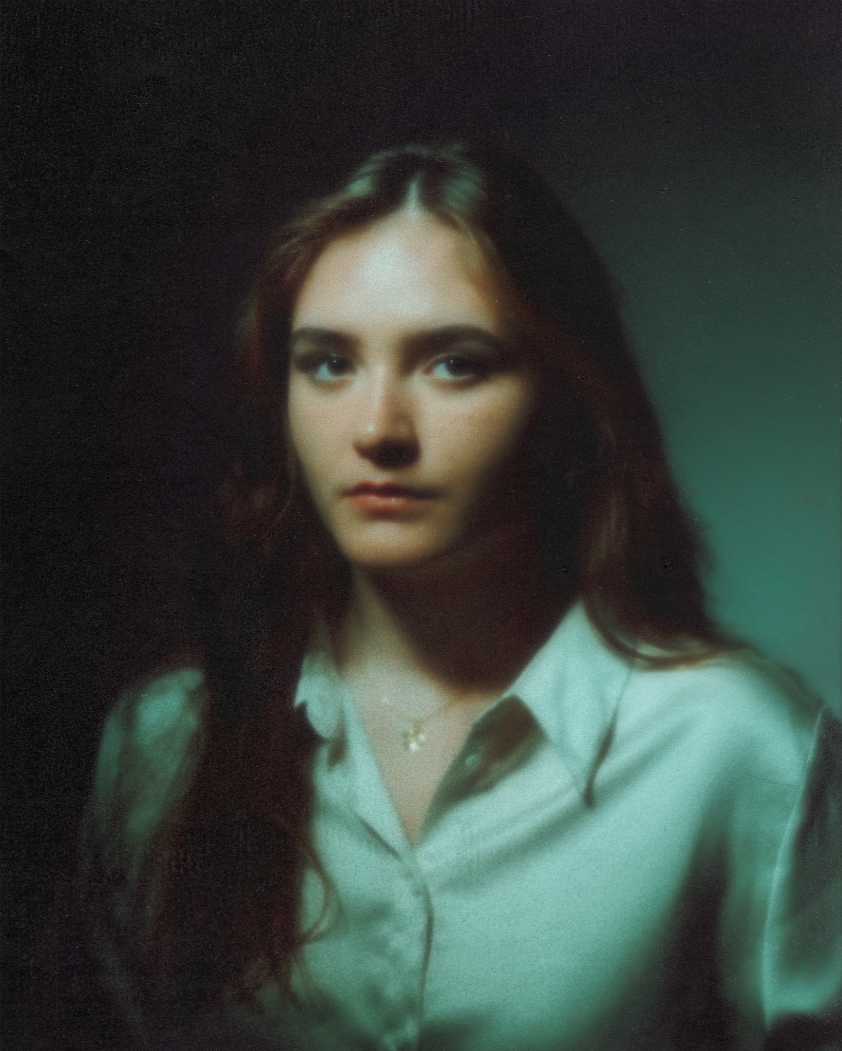 Portret młodej kobiety z długimi, brązowymi włosami, ubranej w satynową koszulę, na ciemnym, rozmytym tle. Delikatne światło podkreśla rysy twarzy i subtelny naszyjnik.