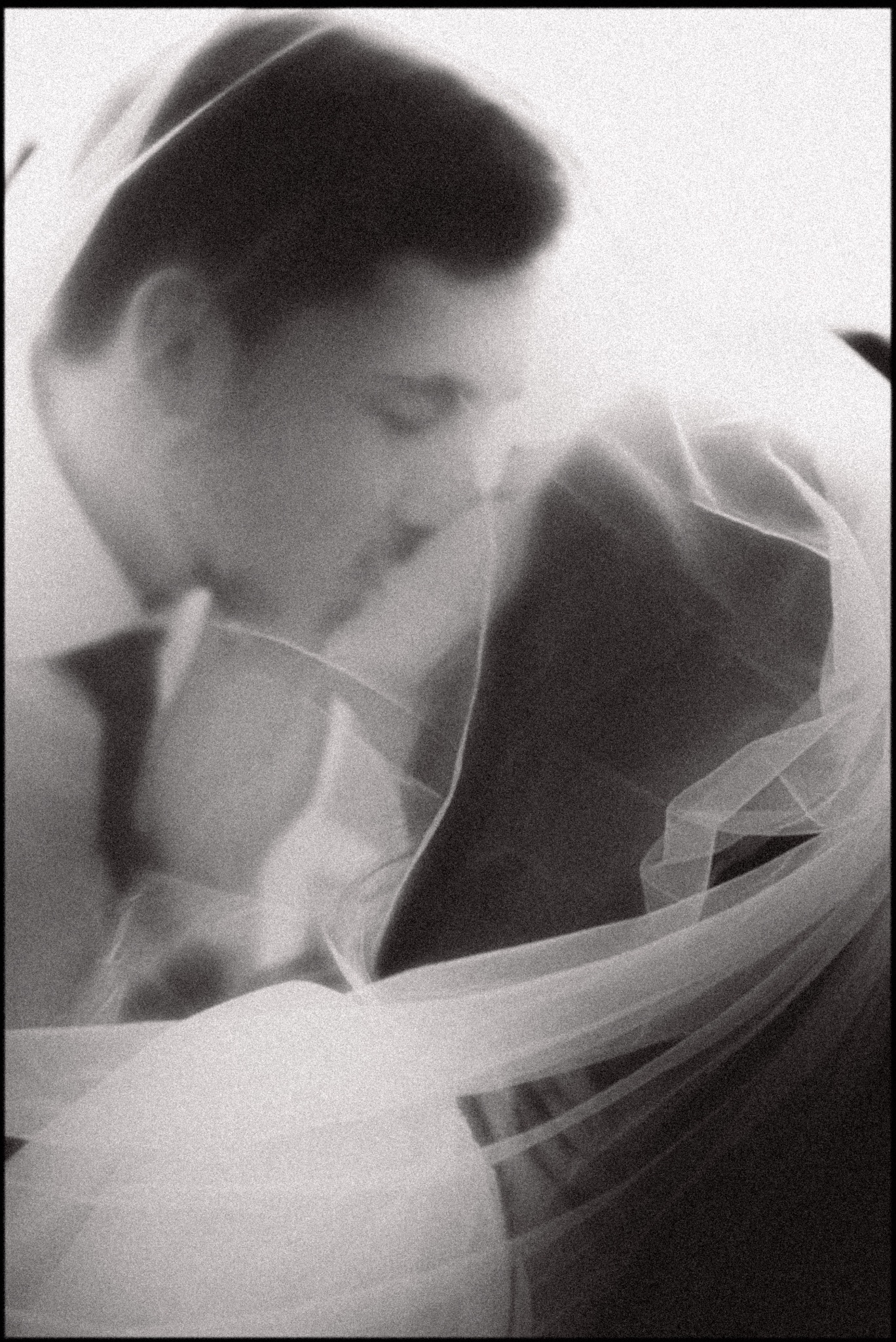 Czarno-białe, artystyczne ujęcie pary młodej w zbliżeniu, z welonem delikatnie okrywającym ich twarze. Subtelne światło i ziarnista faktura dodają intymności.