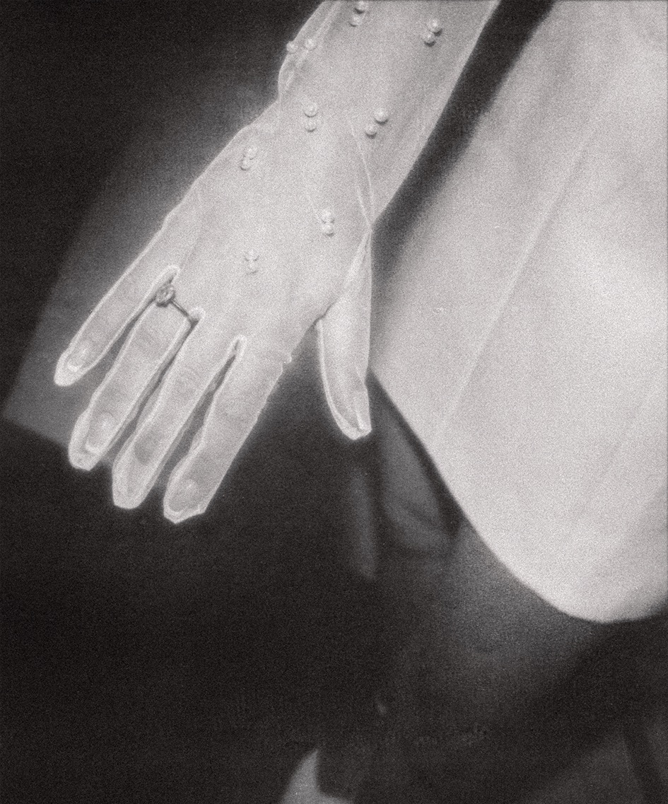 Czarno-białe zbliżenie dłoni w przezroczystej rękawiczce z perełkami, na palcu pierścionek zaręczynowy, delikatne światło, elegancka suknia w tle.