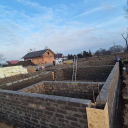 Blacharstwo Dekarstwo,Ciesielstwo usługi ogólnobudowlane.Dach pol - Fundamenty domu z bloczków betonowych, widoczne zbrojenia i szalunki. W tle budynek w trakcie budowy i stos izolacji. Błękitne niebo.