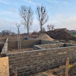 Blacharstwo Dekarstwo,Ciesielstwo usługi ogólnobudowlane.Dach pol - Wznoszone mury fundamentowe z bloczków betonowych na tle ziemi i drzew. Widoczna drabina i deski szalunkowe. Etap budowy domu jednorodzinnego.