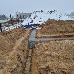 Blacharstwo Dekarstwo,Ciesielstwo usługi ogólnobudowlane.Dach pol - Fundamenty domu w trakcie budowy: wykop zbrojony stalą, częściowo zalany betonem. W tle hałda ziemi i zarys domu w zimowej scenerii.