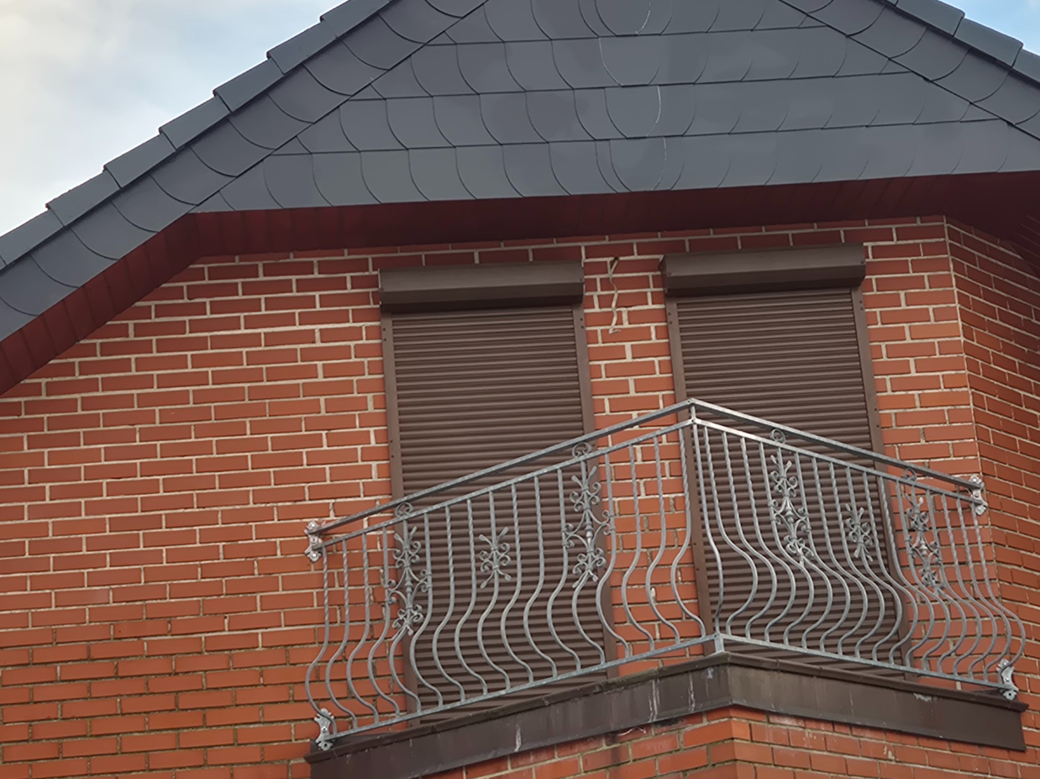 Ceglany budynek z ciemnym dachem krytym łuską, brązowymi roletami i ozdobną, metalową balustradą balkonu. Ujęcie z dołu, widoczne detale wykończenia.