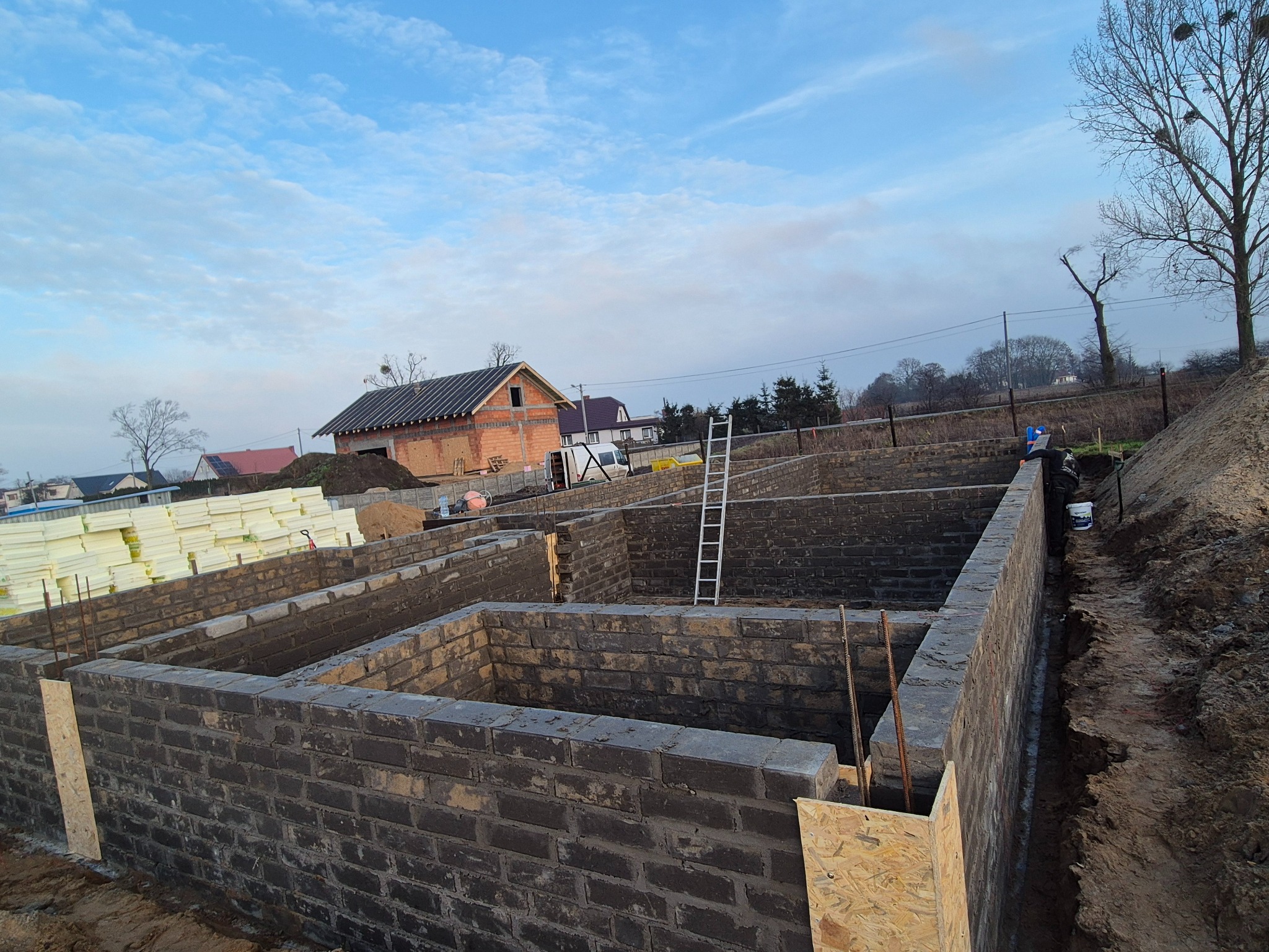 Fundamenty domu z bloczków betonowych, widoczne zbrojenia i szalunki. W tle budynek w trakcie budowy i stos izolacji. Błękitne niebo.