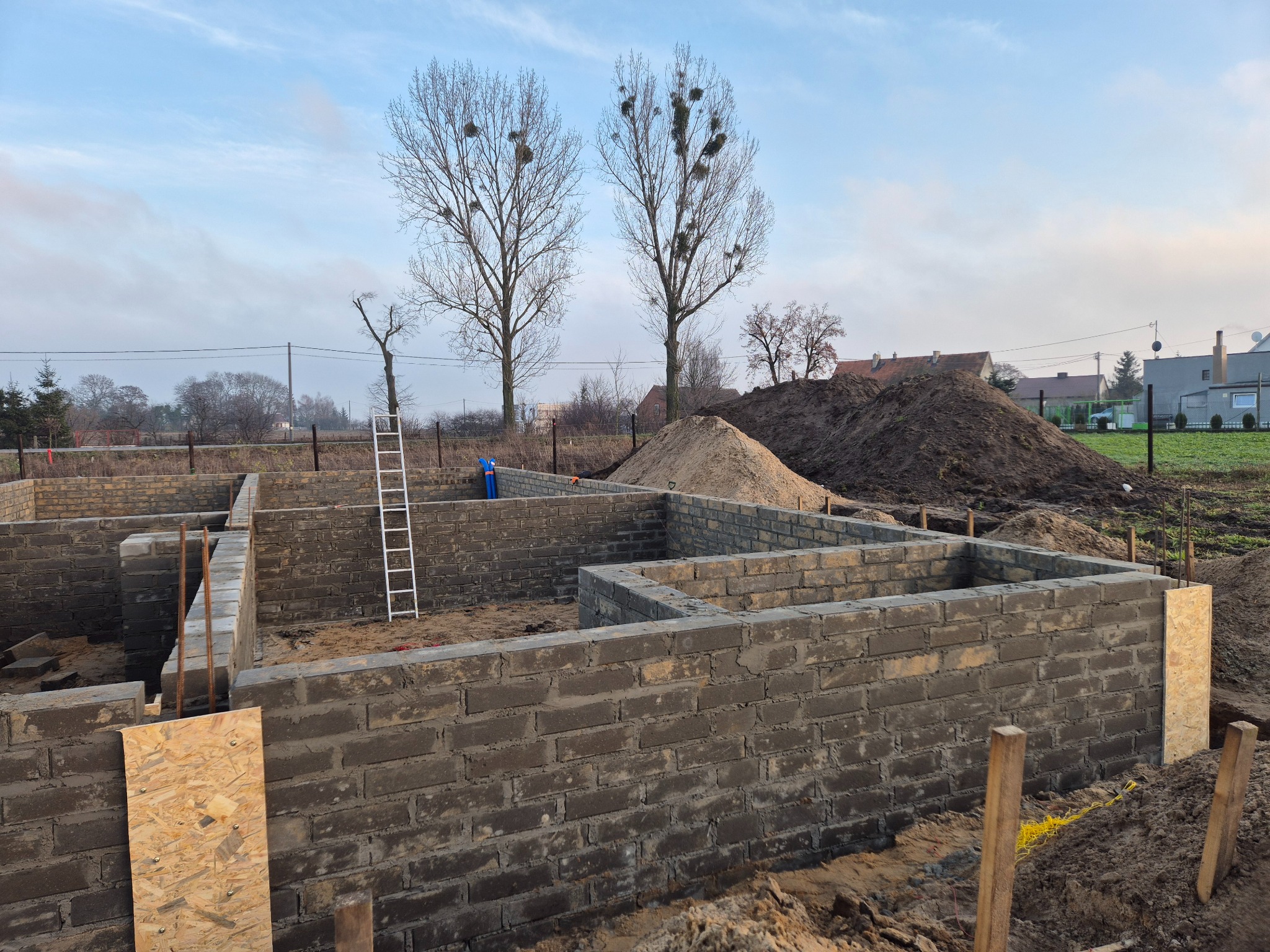Wznoszone mury fundamentowe z bloczków betonowych na tle ziemi i drzew. Widoczna drabina i deski szalunkowe. Etap budowy domu jednorodzinnego.