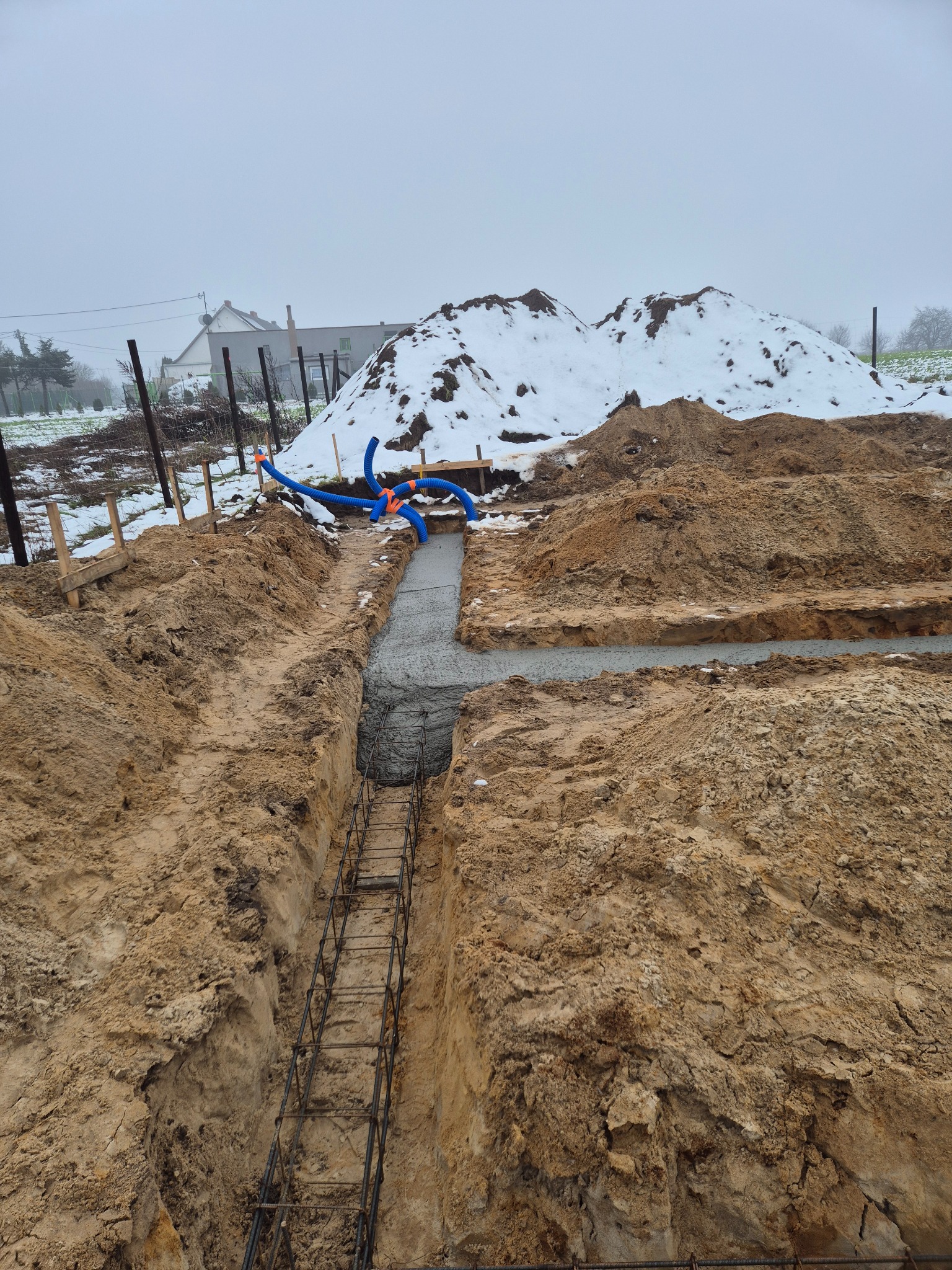 Fundamenty domu w trakcie budowy: wykop zbrojony stalą, częściowo zalany betonem. W tle hałda ziemi i zarys domu w zimowej scenerii.