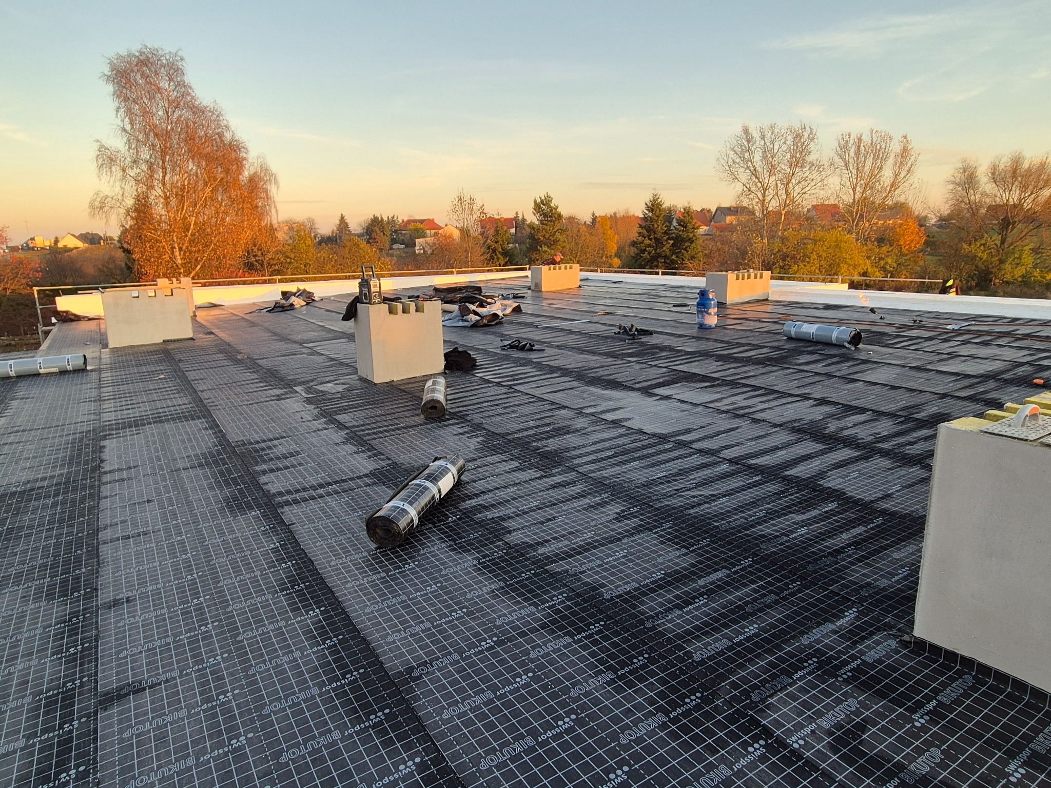 Płaski dach pokryty czarną membraną hydroizolacyjną z widocznymi rolkami materiału, kominami wentylacyjnymi i narzędziami. W tle drzewa i zabudowa mieszkalna o zmierzchu.