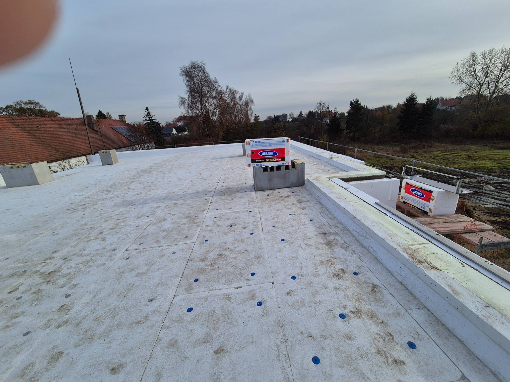 Płaski dach pokryty izolacją termiczną z widocznymi niebieskimi kołkami mocującymi. Na dachu leżą paczki materiału izolacyjnego.