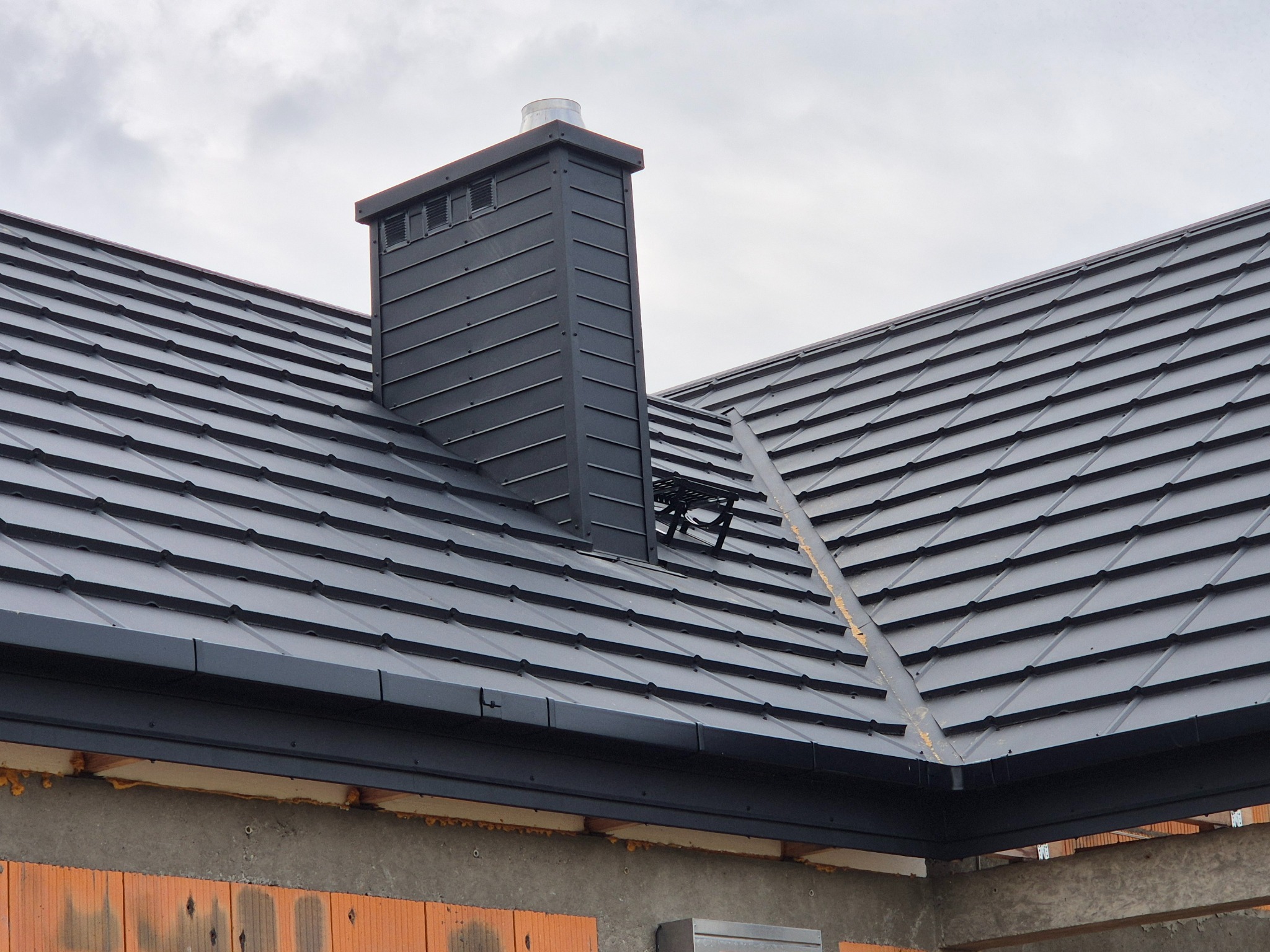 Czarny dach z blachy na domu w budowie, widoczny komin i fragment ściany z cegły. Nowoczesne pokrycie dachowe z blachy trapezowej.