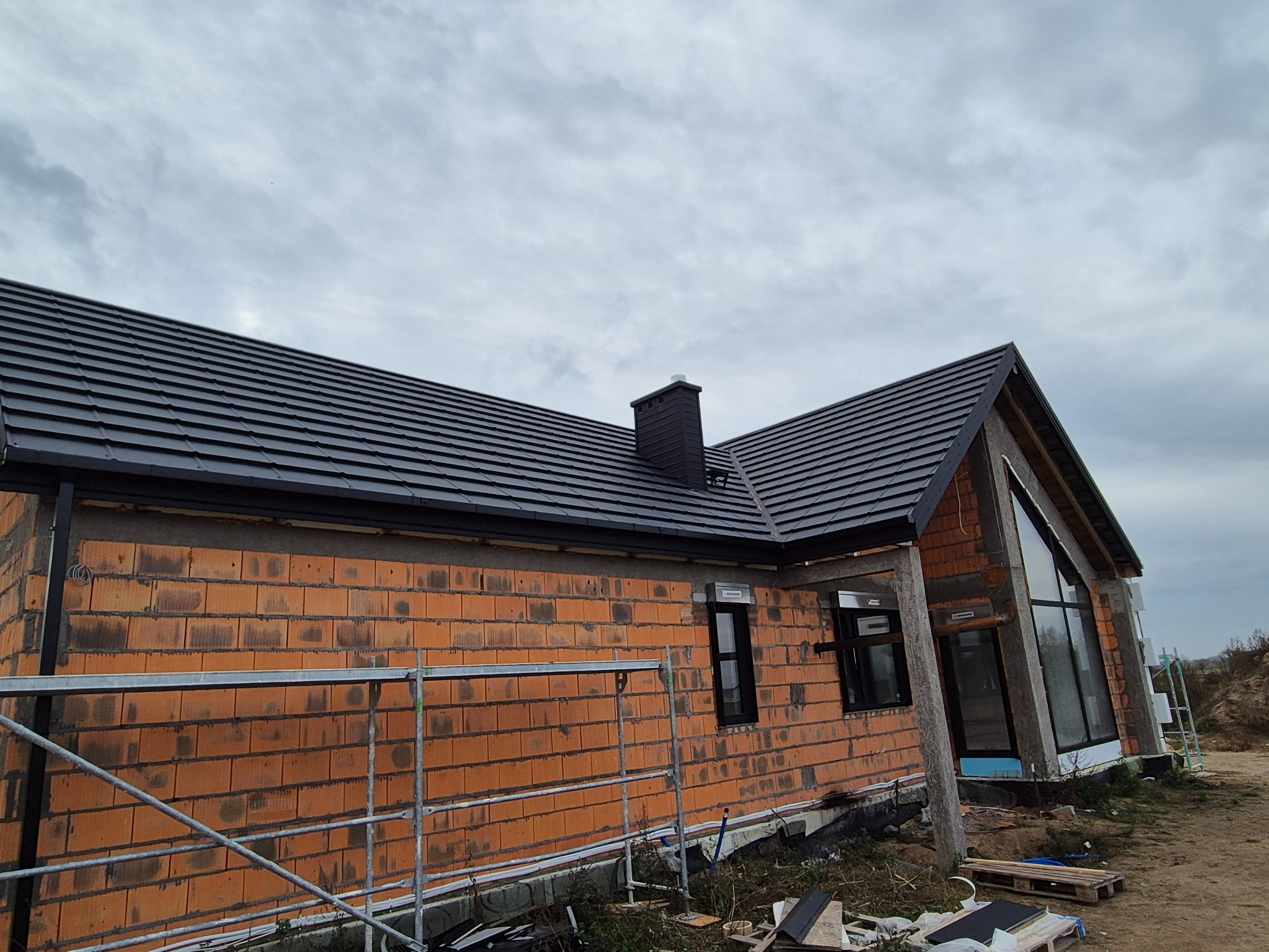 Dom w trakcie budowy z ciemną dachówką i ceglaną elewacją. Widoczne rusztowania i elementy wykończeniowe. Pochmurne niebo w tle.