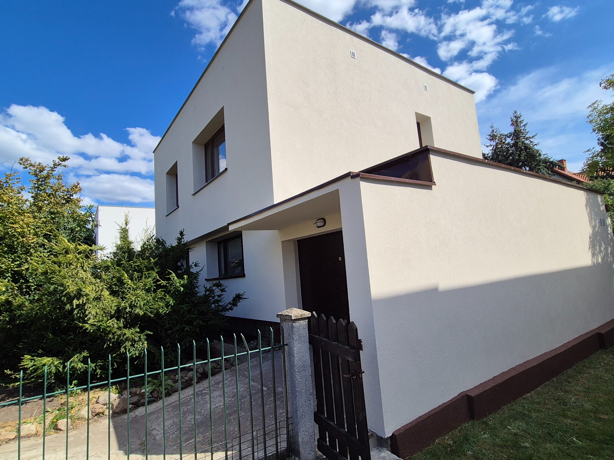 Nowoczesny, otynkowany dom jednorodzinny z płaskim dachem, widziany z poziomu gruntu, z zielonym ogrodzeniem i bujną roślinnością w tle, w słoneczny dzień.