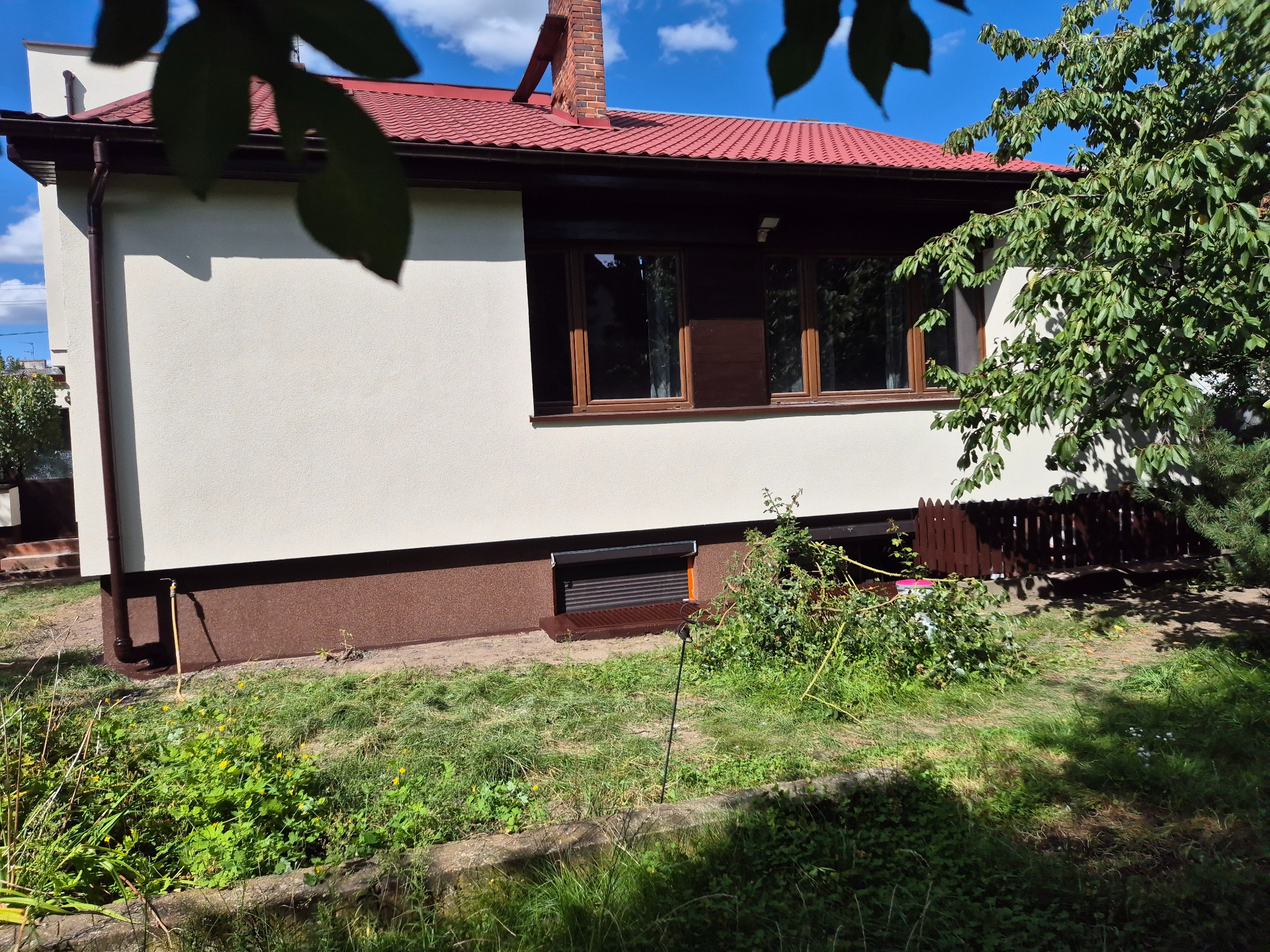 Dom jednorodzinny z czerwoną dachówką, jasną elewacją i brązowymi detalami. Widoczne okna, rynna i fragment ogrodu. Ujęcie w słoneczny dzień.