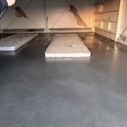 Grzegorz Zadrożny - Błyszcząca, szara posadzka przemysłowa w hali z białymi ścianami. Widoczne fragmenty metalowej konstrukcji dachu i pozostałości po pracach budowlanych.