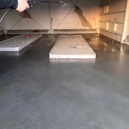 Grzegorz Zadrożny - Aplikacja żywicy na posadzkę przemysłową w hali. Widoczne pozostałości po wcześniejszych pracach i osoba rozpylająca preparat. Błyszcząca, szara powierzchnia.