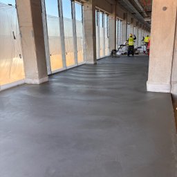 Grzegorz Zadrożny - Wylewka posadzki przemysłowej w surowym wnętrzu budynku. Dwóch pracowników w żółtych kamizelkach używa zacieraczek mechanicznych. Okna zabezpieczone folią.