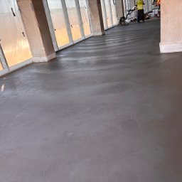 Grzegorz Zadrożny - Wylewka posadzki przemysłowej w trakcie realizacji. Widoczna maszyna do zacierania betonu i pracownik w odblaskowej kamizelce. Świeża, szara powierzchnia posadzki.