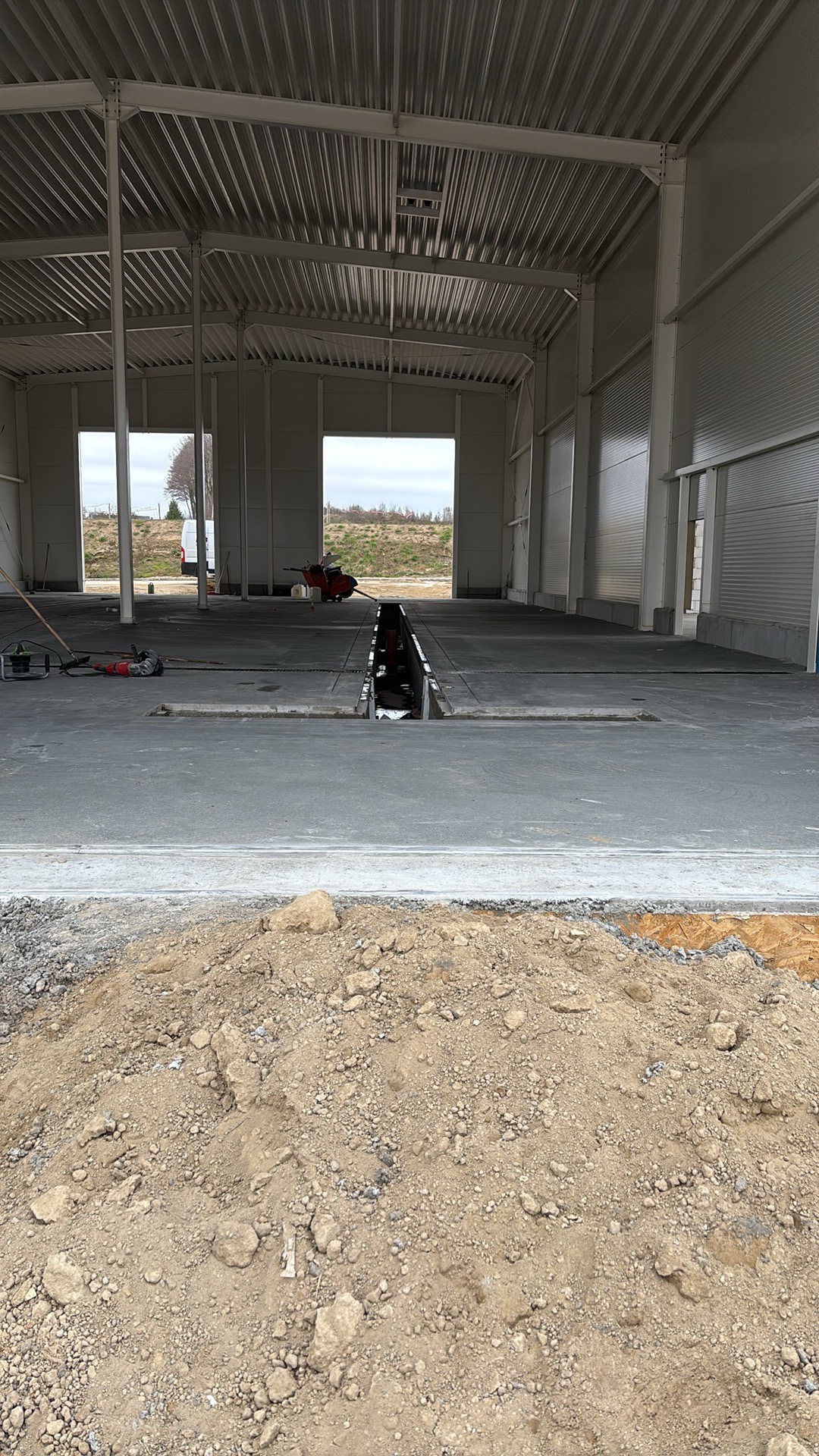 Wnętrze hali w trakcie budowy z wylewką posadzki przemysłowej. Widoczne narzędzia i wycięty fragment posadzki. Na pierwszym planie hałda ziemi.