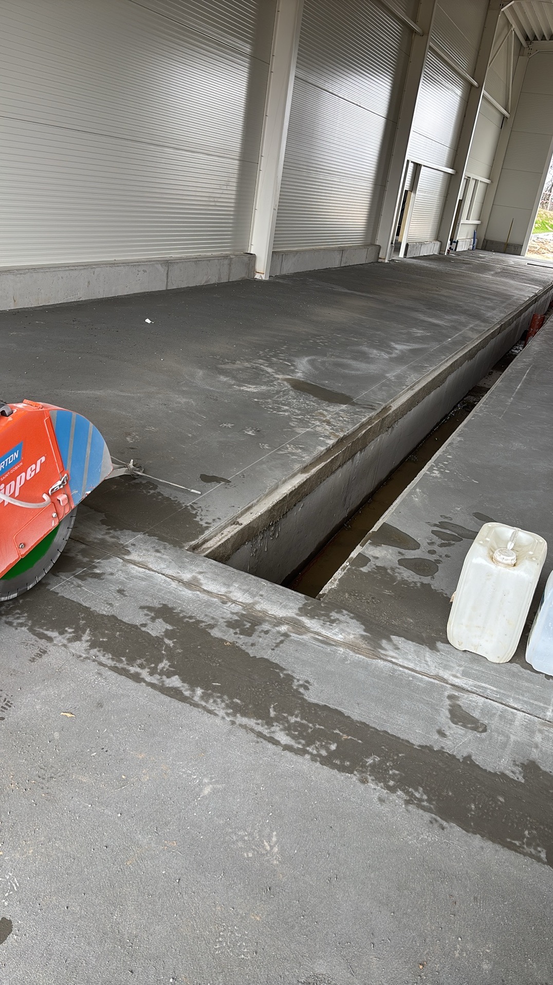 Cięcie dylatacji w posadzce przemysłowej z użyciem przecinarki, widoczne białe kanistry z płynem chłodzącym. Hala z blachy trapezowej w tle.