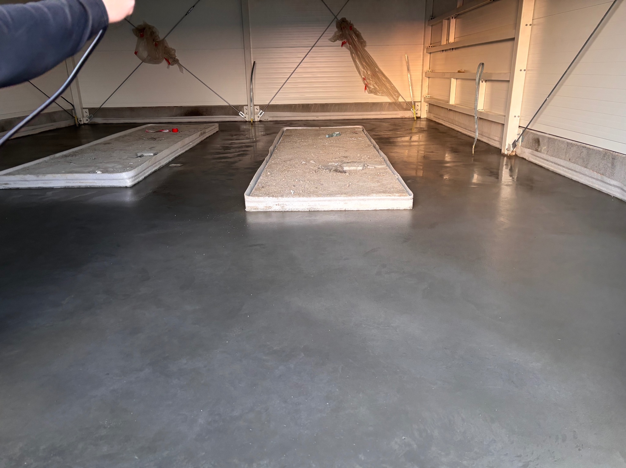 Błyszcząca, szara posadzka przemysłowa w hali z białymi ścianami. Widoczne fragmenty metalowej konstrukcji dachu i pozostałości po pracach budowlanych.