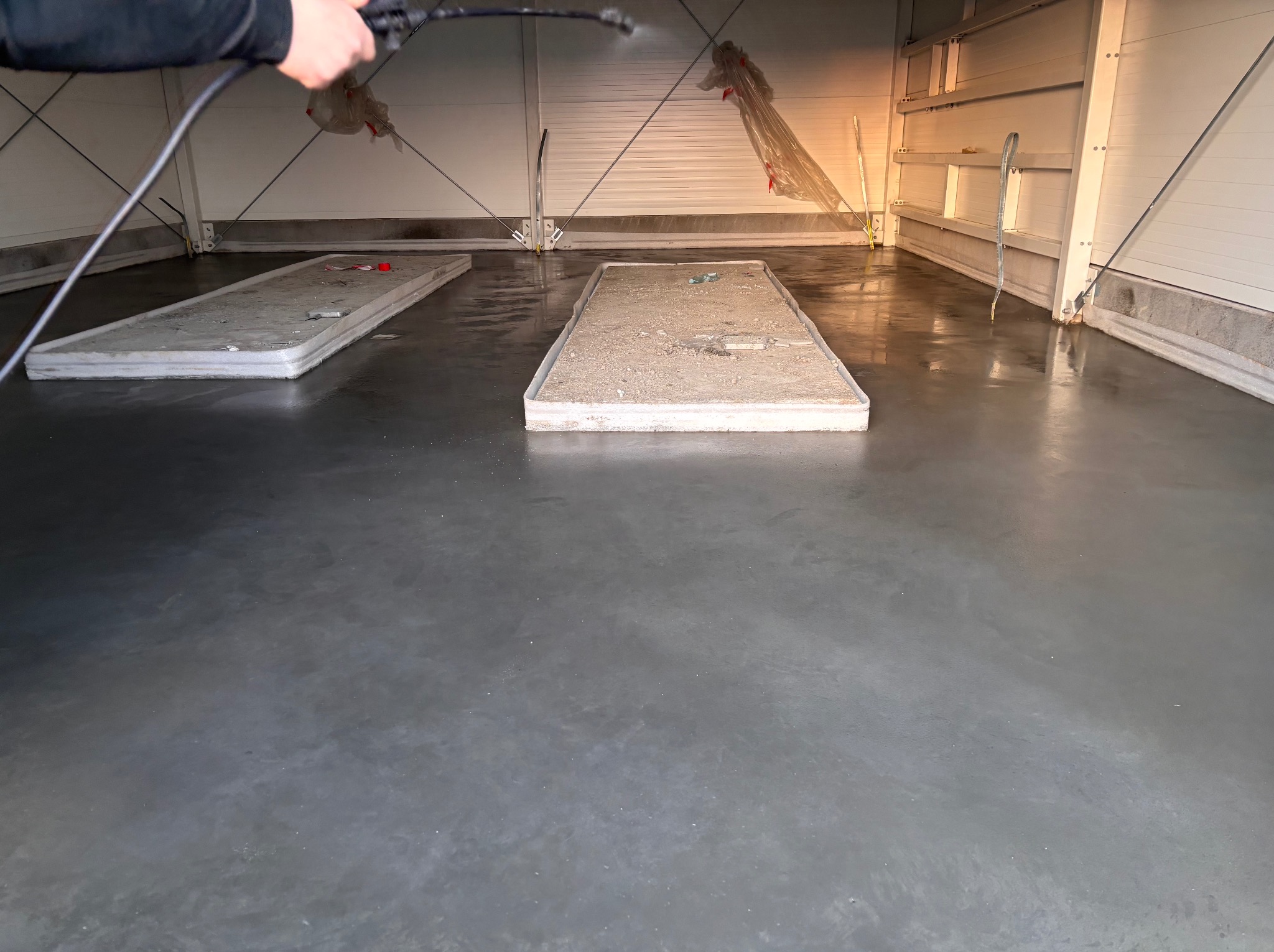 Aplikacja żywicy na posadzkę przemysłową w hali. Widoczne pozostałości po wcześniejszych pracach i osoba rozpylająca preparat. Błyszcząca, szara powierzchnia.