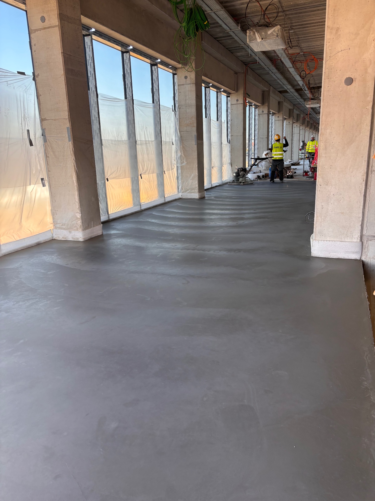 Wylewka posadzki przemysłowej w surowym wnętrzu budynku. Dwóch pracowników w żółtych kamizelkach używa zacieraczek mechanicznych. Okna zabezpieczone folią.