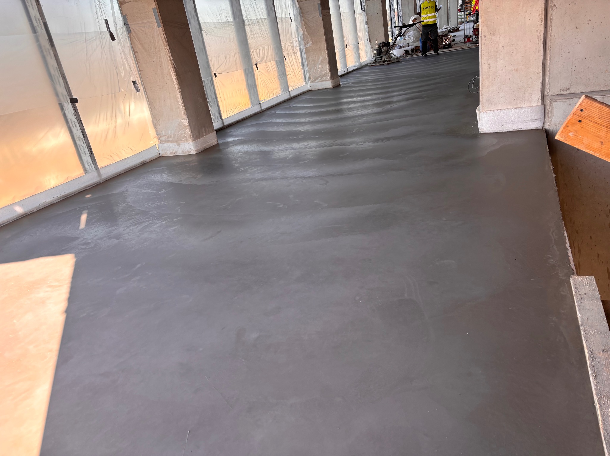 Wylewka posadzki przemysłowej w trakcie realizacji. Widoczna maszyna do zacierania betonu i pracownik w odblaskowej kamizelce. Świeża, szara powierzchnia posadzki.