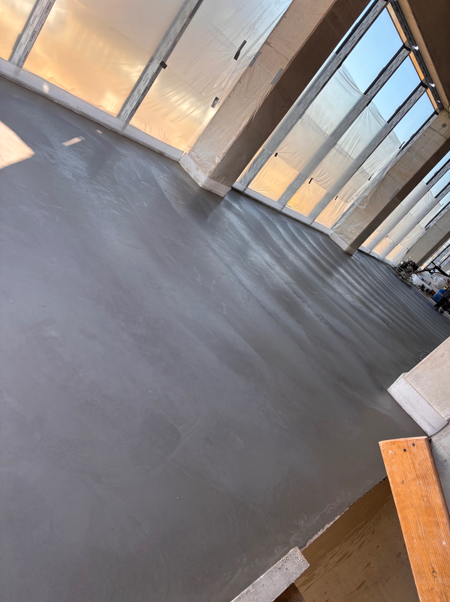 Świeżo wylana, gładka posadzka przemysłowa w jasnym wnętrzu z oknami zabezpieczonymi folią. Widoczne narzędzia i fragment deski szalunkowej.