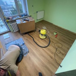 GO PROJECT - Układanie paneli podłogowych w pokoju o zielonych ścianach. Widoczny odkurzacz przemysłowy, narzędzia i fragment osoby podczas pracy. Ujęcie z góry.