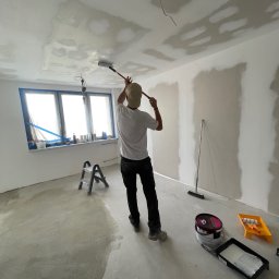 GO PROJECT - Malowanie sufitu wałkiem w pomieszczeniu z oknem i przygotowanymi ścianami. Widoczne narzędzia malarskie i drabina. Praca w toku, sufit częściowo pomalowany.