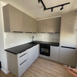 GO PROJECT - Nowoczesna kuchnia w odcieniach szarości z czarnym blatem i minimalistycznymi uchwytami, oświetlona reflektorami sufitowymi. Widok na okno z żaluzjami.