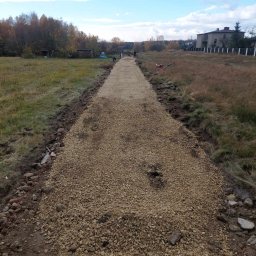 AKH INVEST - Utwardzona droga gruntowa z jasnym kruszywem, widoczne ślady po bokach, w tle drzewa i budynek. Prace ziemne w toku, zarysowany kształt drogi.