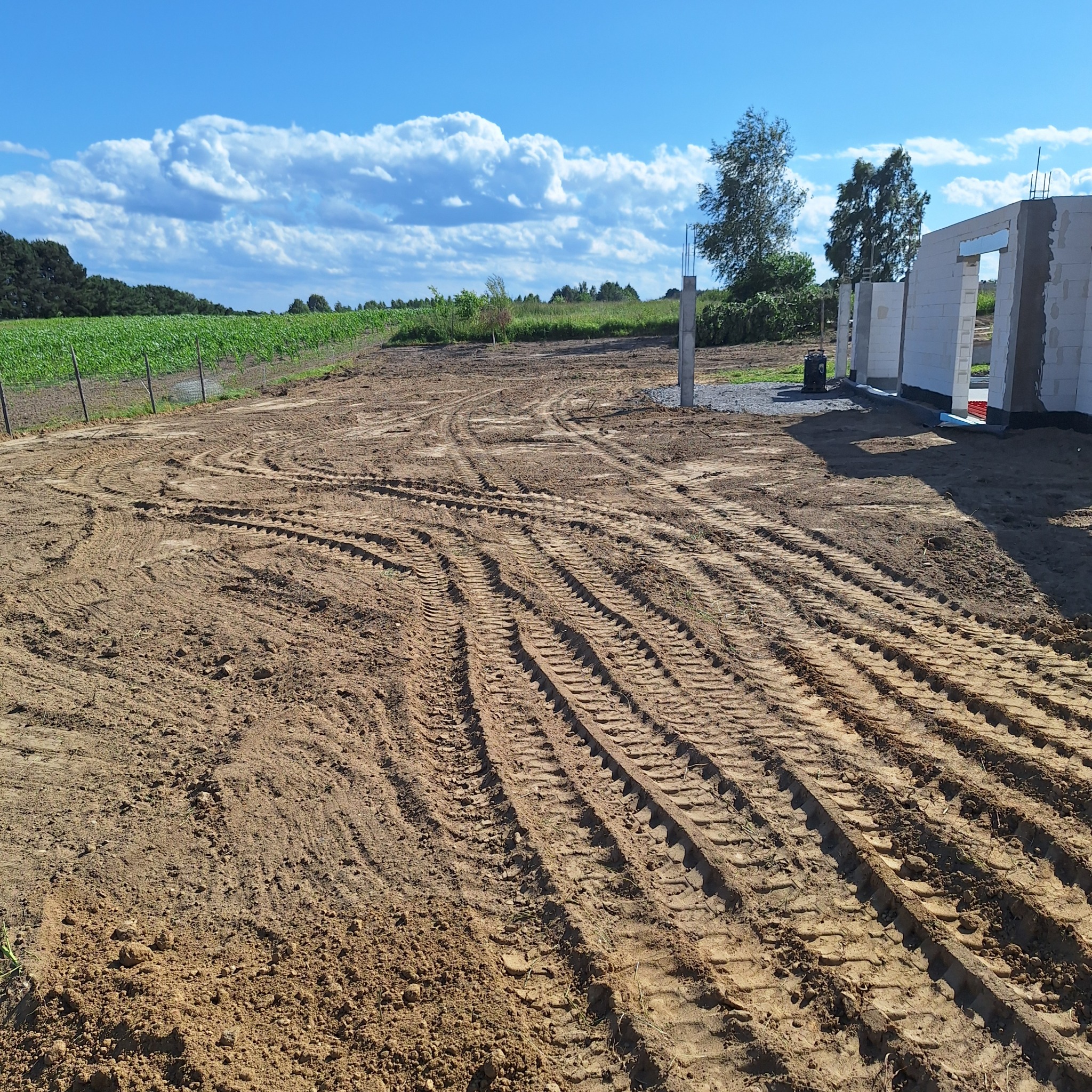 Ślady opon na ziemi po niwelacji terenu pod budowę domu jednorodzinnego. W tle widoczne fundamenty i zielone pola uprawne pod błękitnym niebem.