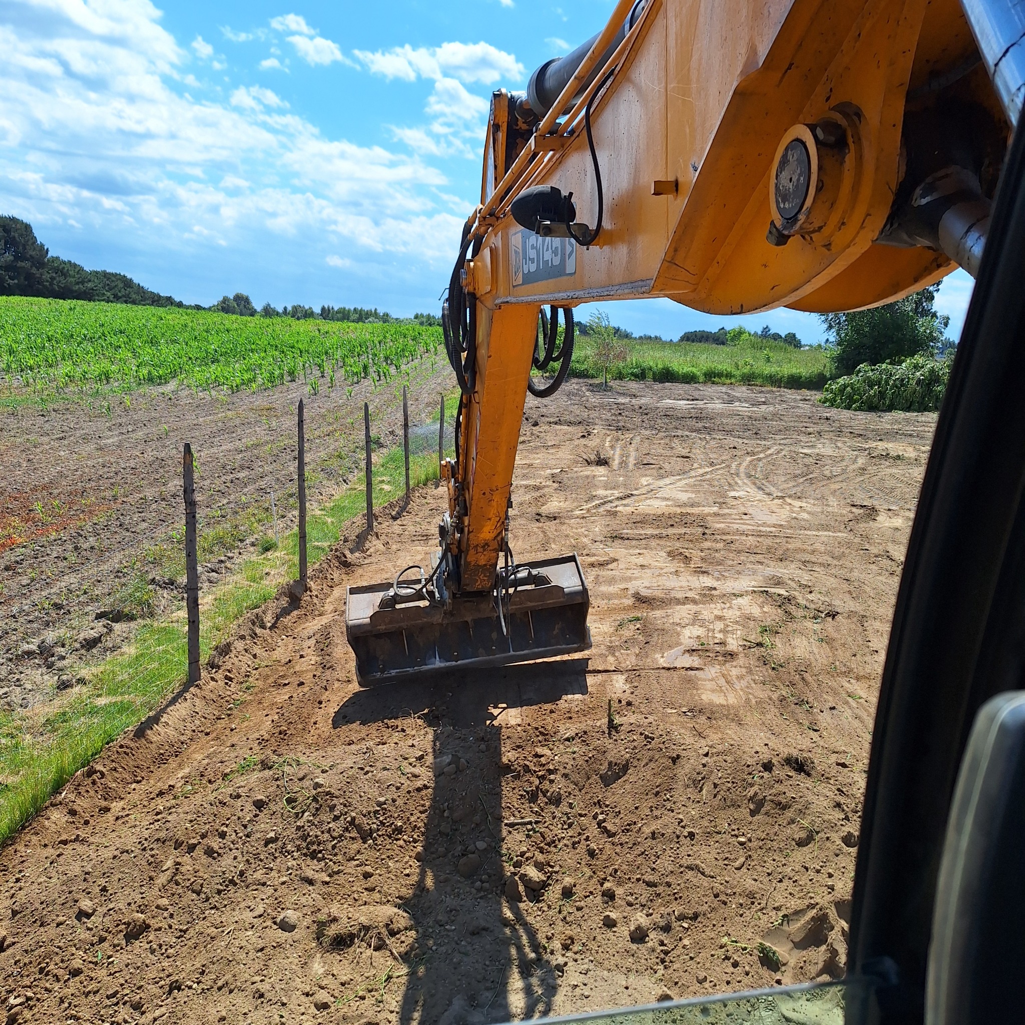 Żółta koparka JS145 wyrównuje teren wzdłuż ogrodzenia na tle zielonego pola i błękitnego nieba. Widok z kabiny operatora.