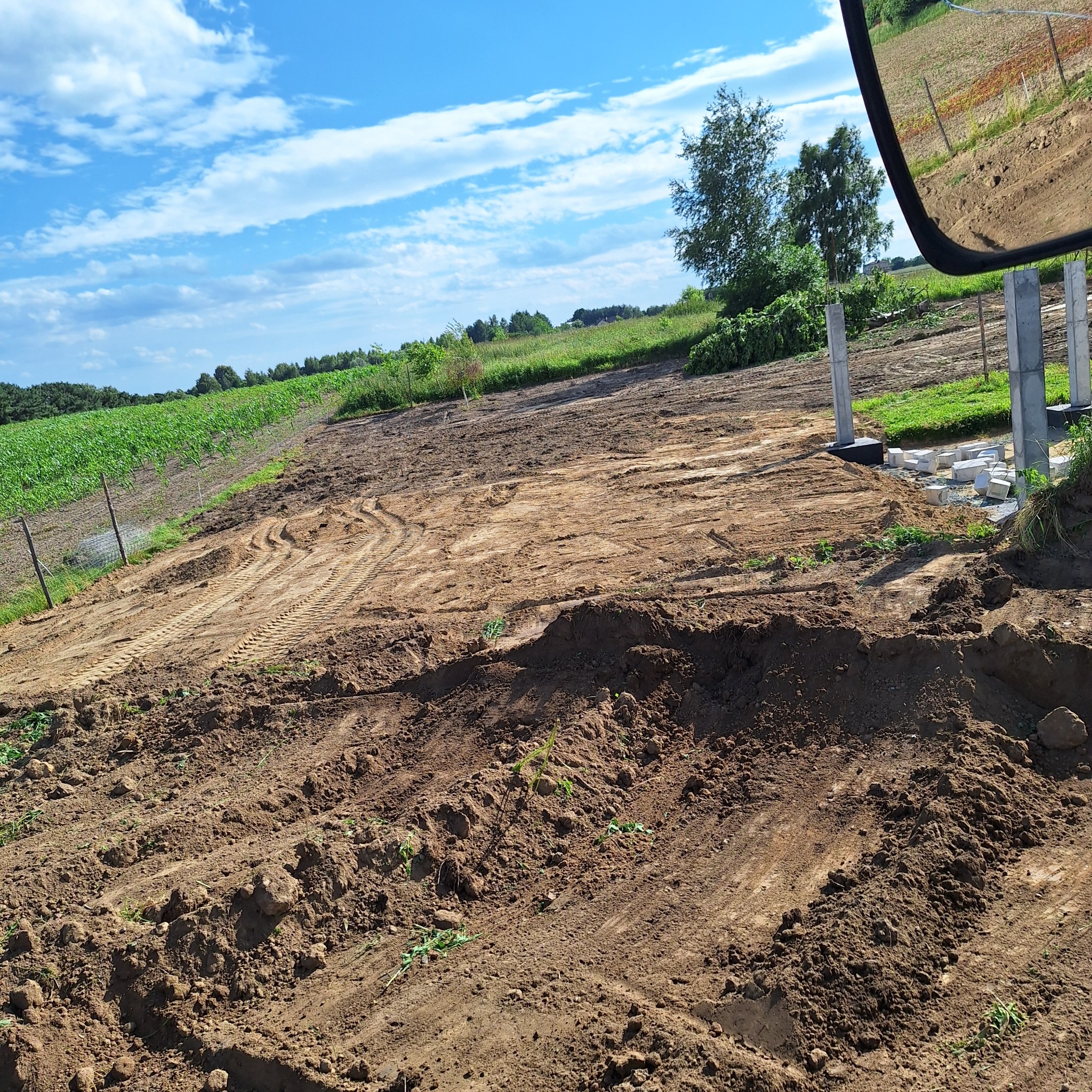 Przygotowany teren pod budowę z widocznymi śladami opon i betonowymi słupami w tle, częściowo odbitymi w lusterku pojazdu. Błękitne niebo z chmurami.