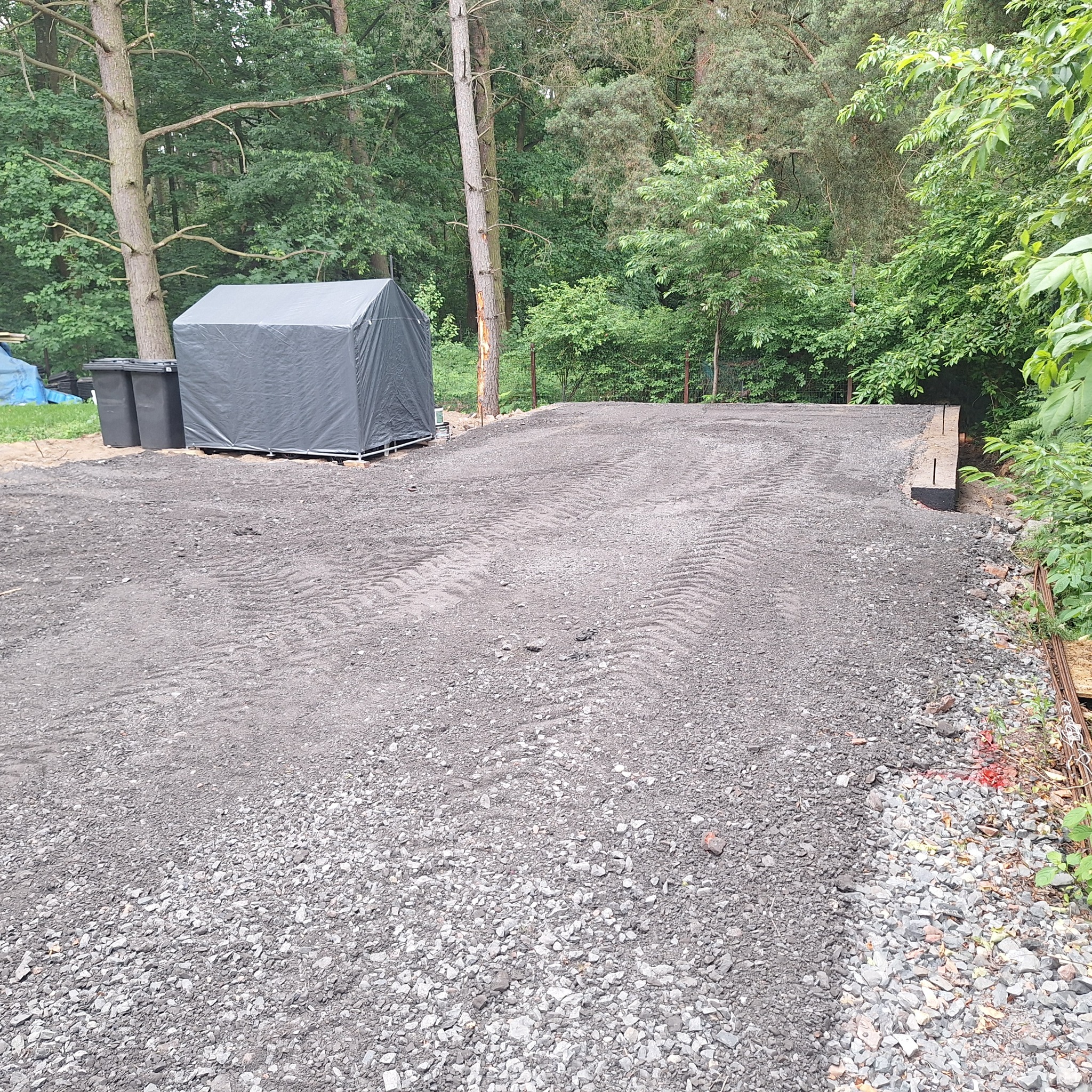 Utwardzony teren z widocznymi śladami opon, obok zadaszona konstrukcja i drzewa w tle. Widoczne elementy infrastruktury i roślinność.